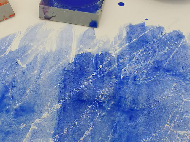 Blaue Farbe übermalt Wachsspur auf einem Blatt Papier.