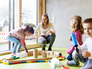 Eine Erzieherin sitzt mit einigen Kindergartenkindern in der Kita auf dem Boden der Gruppenraums. Sie pusten Kugeln durch einen Parcours.