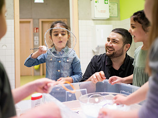 Eine Erzieherin sitzt mit einigen Kindergartenkindern in der Kita auf dem Boden der Gruppenraums. Sie pusten Kugeln durch einen Parcours.